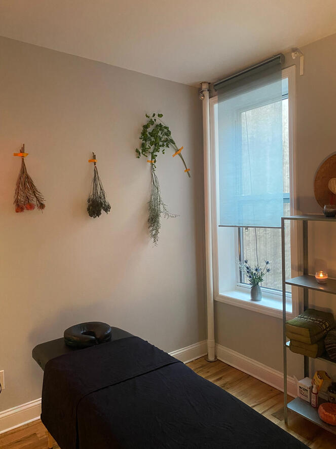 Photo of the massage table in the middle of the session room. There is a vase of dried flowers in the window and dried flowers on the wall.